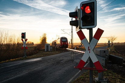 Ein Zug durchquert einen Bahnübergang. Der Bahnübergang ist mit einer Schranke gesichert. Vor der Schranke steht eine rote Ampel.