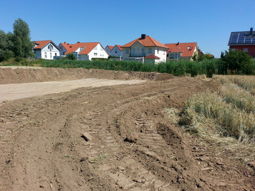 Auf dem Bild eine Geländeauffüllung vor dem Hintergrund eines angrenzenden Baugebiets zu sehen.